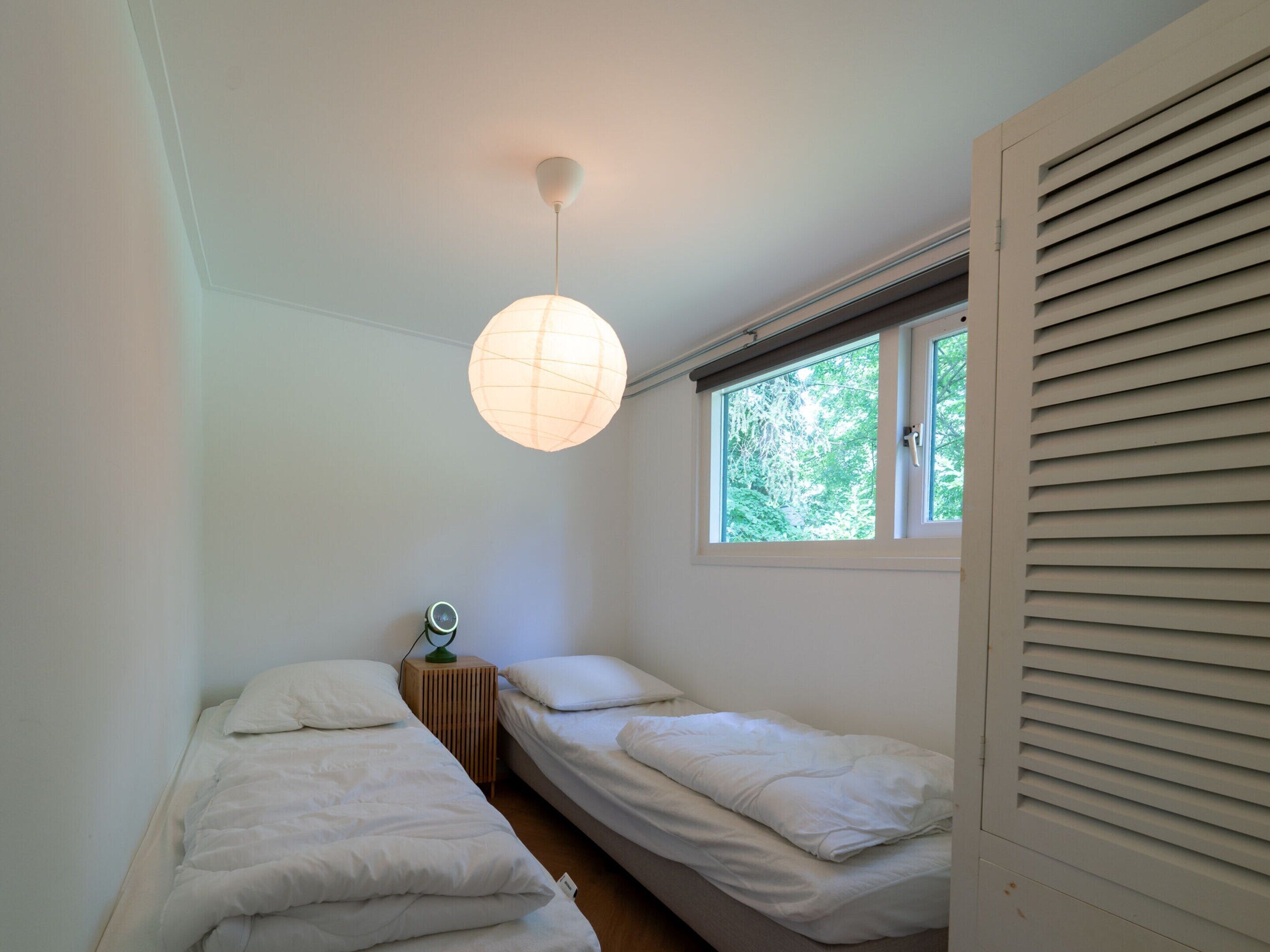 A small, single-room bedroom with two beds, a window with blinds, a hanging light fixture, and a small wooden nightstand with a lamp.