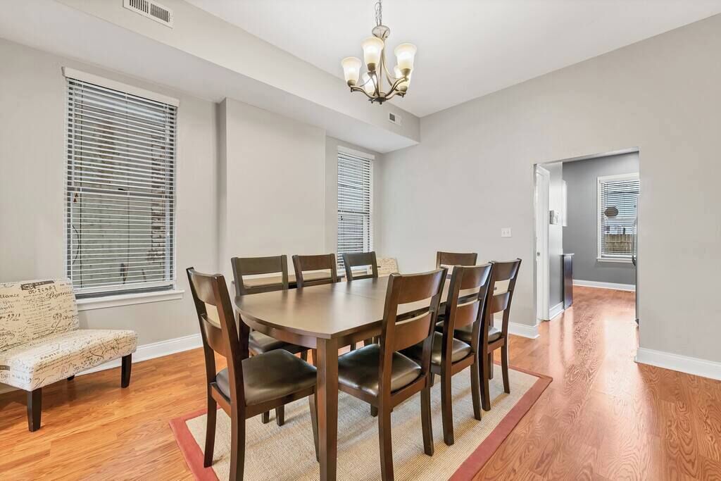 A dining room with a wooden table and chairs, a chandelier, and large windows with blinds.