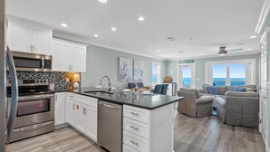 Una cocina moderna con armarios blancos, electrodomésticos de acero inoxidable y una isla grande. Hay una zona de estar con vistas al océano.