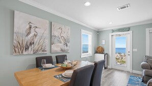 Un comedor con una mesa de madera dispuesta para cuatro personas, rodeada de sillas. Hay dos cuadros de aves colgados en la pared, una puerta con vistas a la playa y un mueble pequeño con un objeto decorativo.