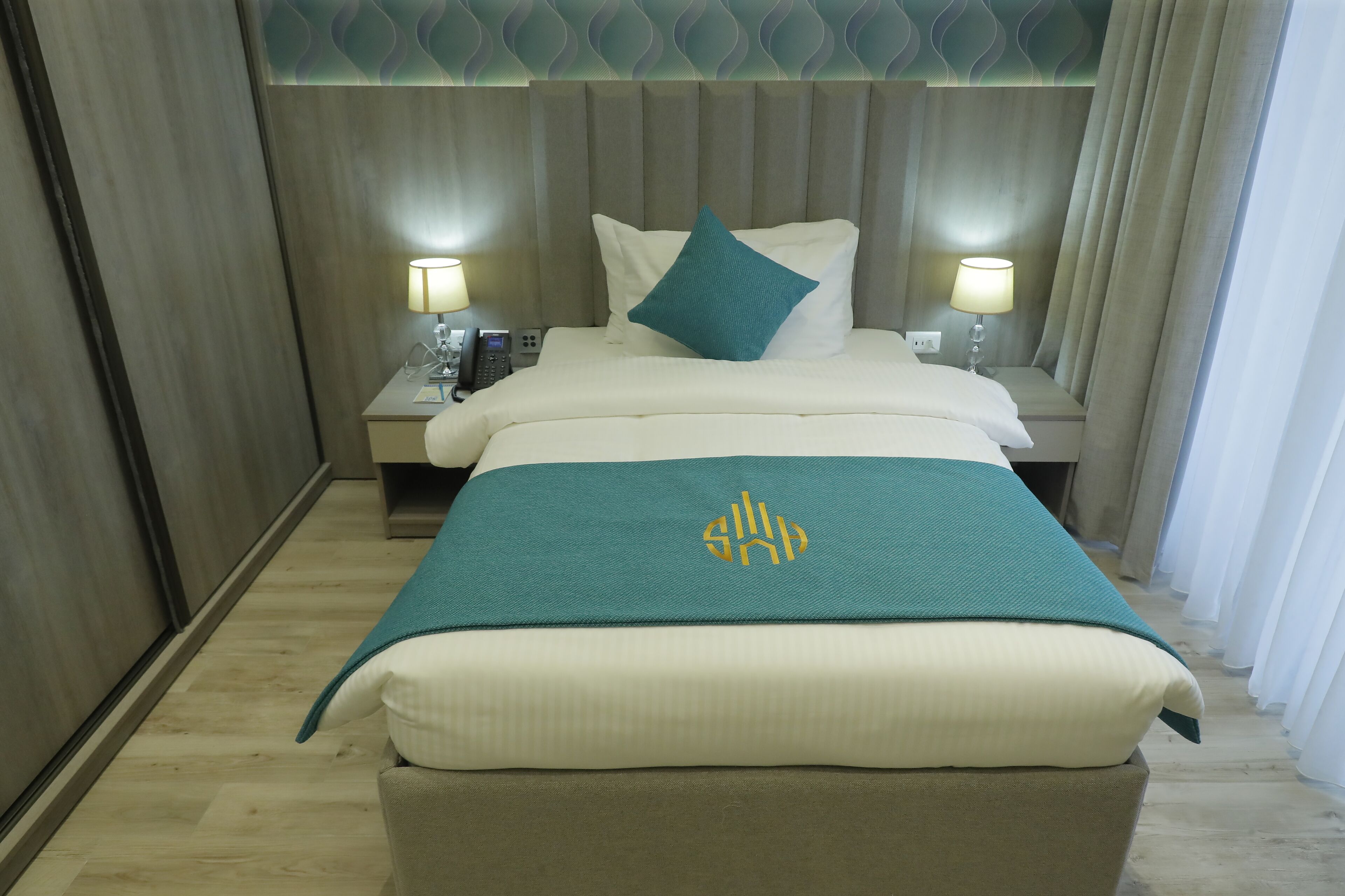 Une chambre d’hôtel comprenant un lit, deux tables de chevet avec des lampes, une petite table et un plaid vert orné d’un logo.
