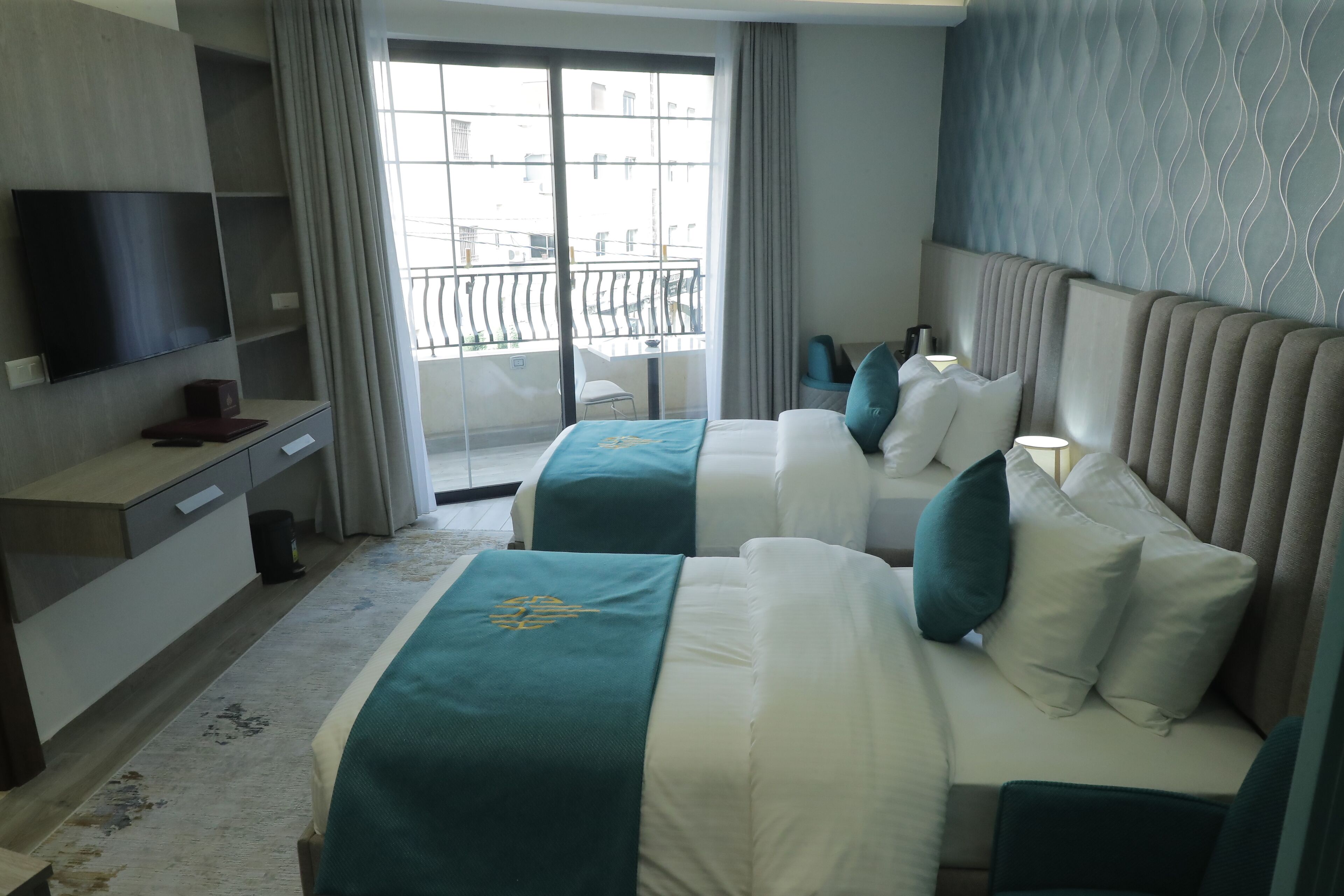 A hotel room with two beds, a TV, a desk, and a balcony with a railing.