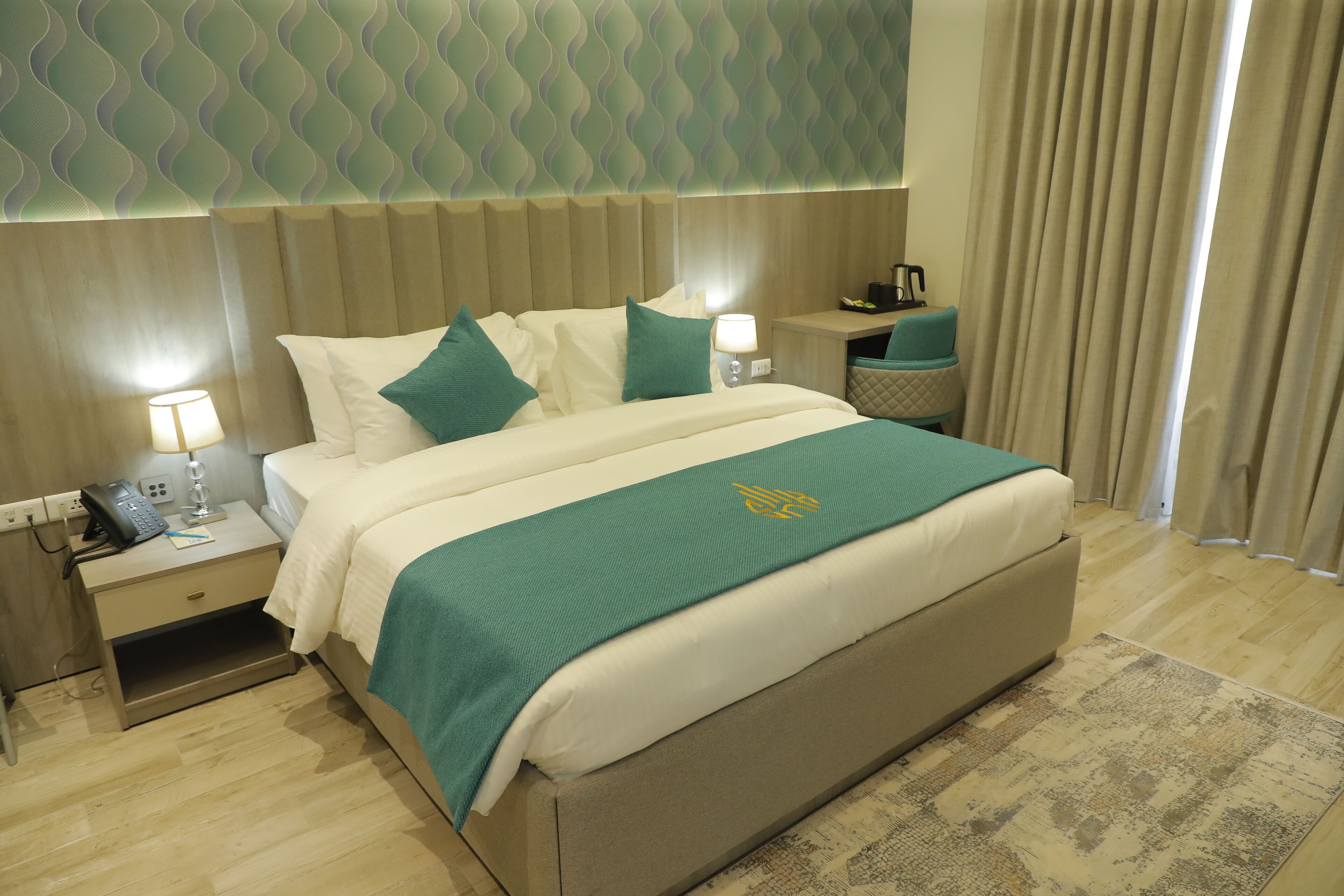 Una camera d'albergo con un letto grande, un comodino con un telefono, una lampada e un tavolino con una tazza e un libro.