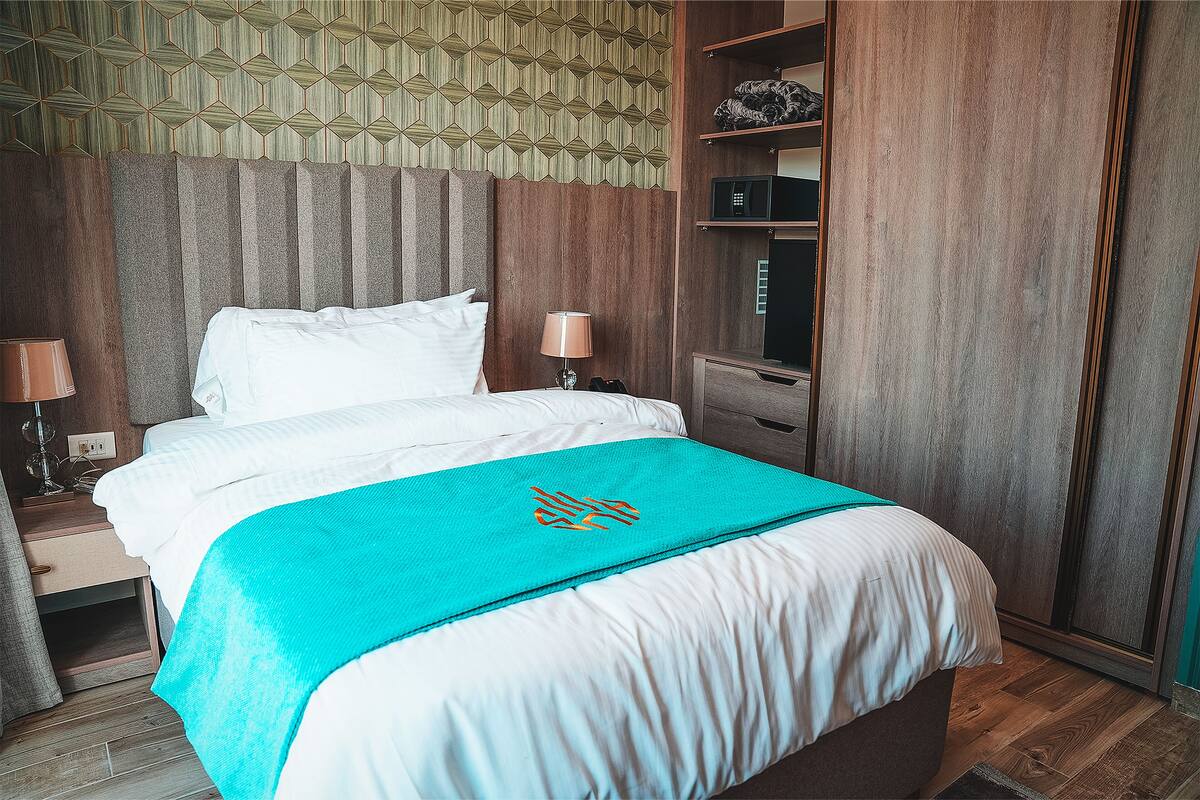 A neatly made bed with a teal throw, a wooden headboard, and a nightstand with a lamp.