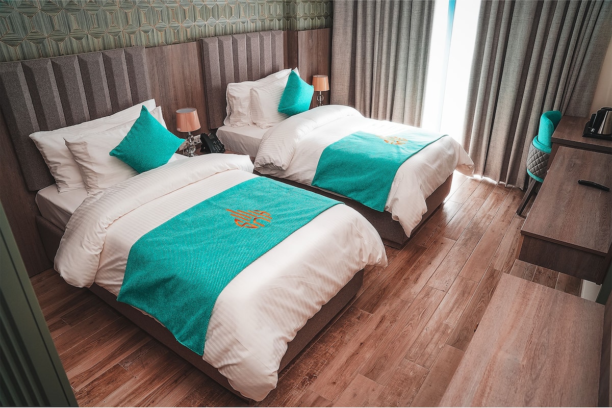 A hotel room with two beds, wooden flooring, a patterned wall, and a view of the outdoors.