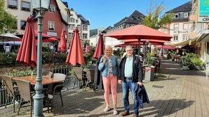 A couple standing in a cobblestone square with outdoor seating and red umbrellas. - Holiday Apartment Magnolia (Ockfen)