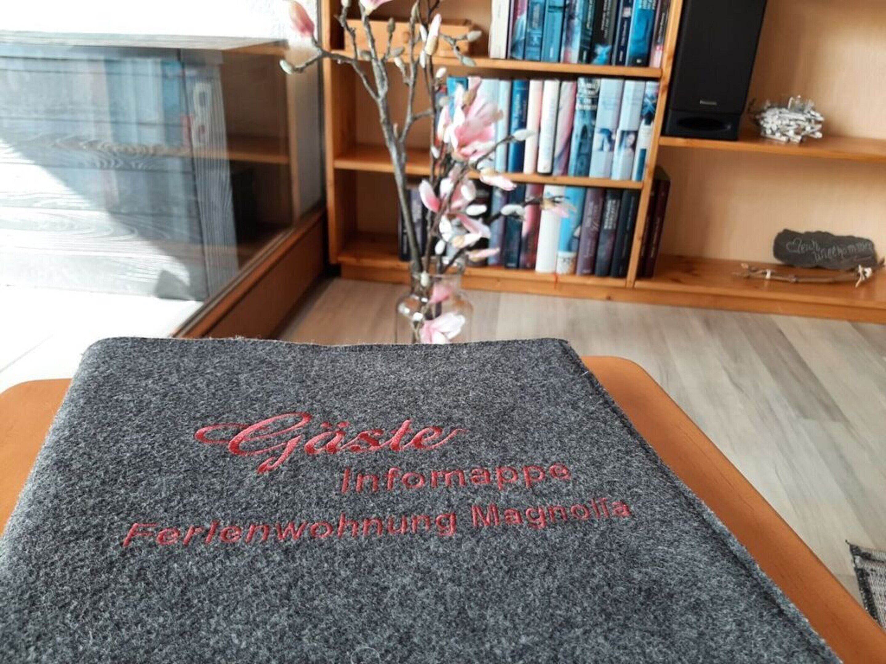 A grey carpet with embroidered text in a room with a wooden bookshelf, a vase with flowers, and a window in the background.