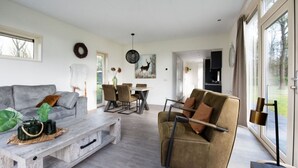 A modern living room with a grey sofa, a wooden coffee table, and a brown armchair. There is a dining area with a white table and chairs, and a kitchen visible through an open door.