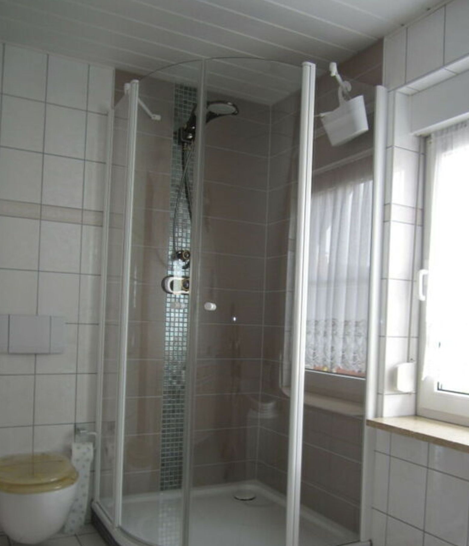 Eine begehbare Dusche mit Glastür, gefliesten Wänden und einem Fenster mit Vorhängen.