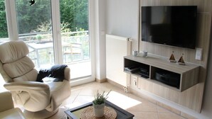 A living room with a large TV, a comfortable chair, a coffee table with a plant, and a view of the outdoors through a sliding glass door. - Apartment Near the Beach With sun Balcony (Boltenhagen)