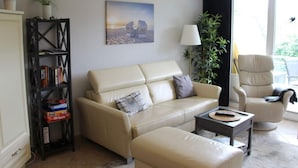 A living room with a beige sofa, a matching armchair, a coffee table, a bookshelf, and a painting on the wall. - Apartment Near the Beach With sun Balcony (Boltenhagen)