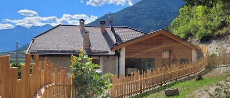 A wooden cabin with a sloped roof, surrounded by a wooden fence, set against a mountainous backdrop.