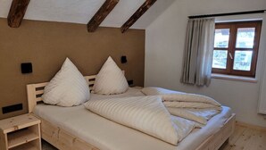 Un dormitorio con cama de madera, dos almohadas, una mesita de noche, una ventana con cortinas y un viguelo de madera en el techo.
