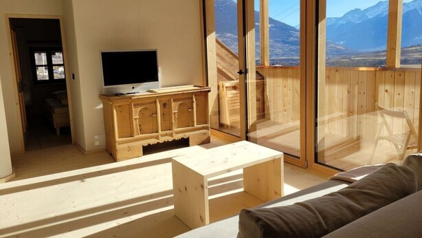 Una sala de estar moderna con un televisor sobre un mueble de madera, un sofá y vistas a las montañas a través de amplios ventanales.