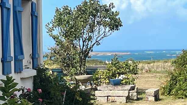 Un jardín con vista al mar, bancos de piedra y un edificio con contraventanas azules.