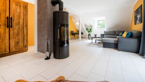 A modern living room with a central black wood-burning stove, a wooden wardrobe, a grey sofa, and a dining area with a table and chairs. - Maarchalet Modern Retreat (Daun)