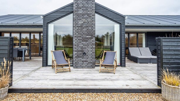 Een modern huis met een centraal schoorsteen, grote glasramen en een houten terras met twee loungestoelen.