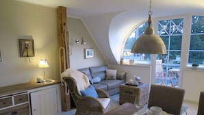 A cozy loft-style living space with a dining area, a sofa, a wicker side table, and a large window with a view.