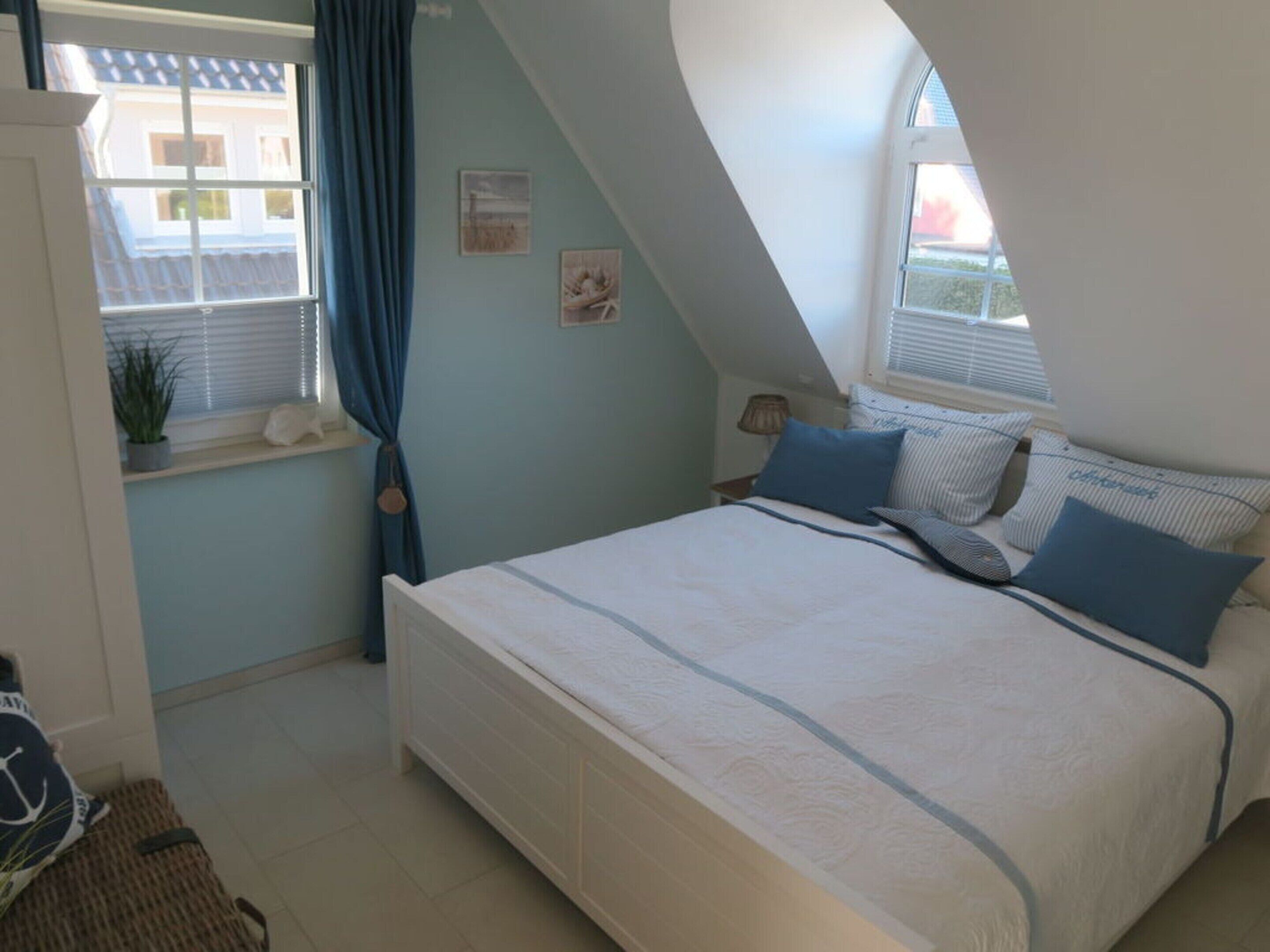 A bedroom with a bed, a window with curtains, a small table with a lamp, and a wall with framed pictures.