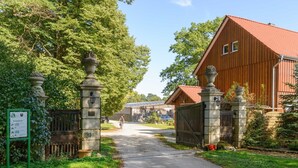 Un chemin de campagne mène à une grange en bois dont le toit est rouge, entourée d’une végétation luxuriante et de piliers en pierre.