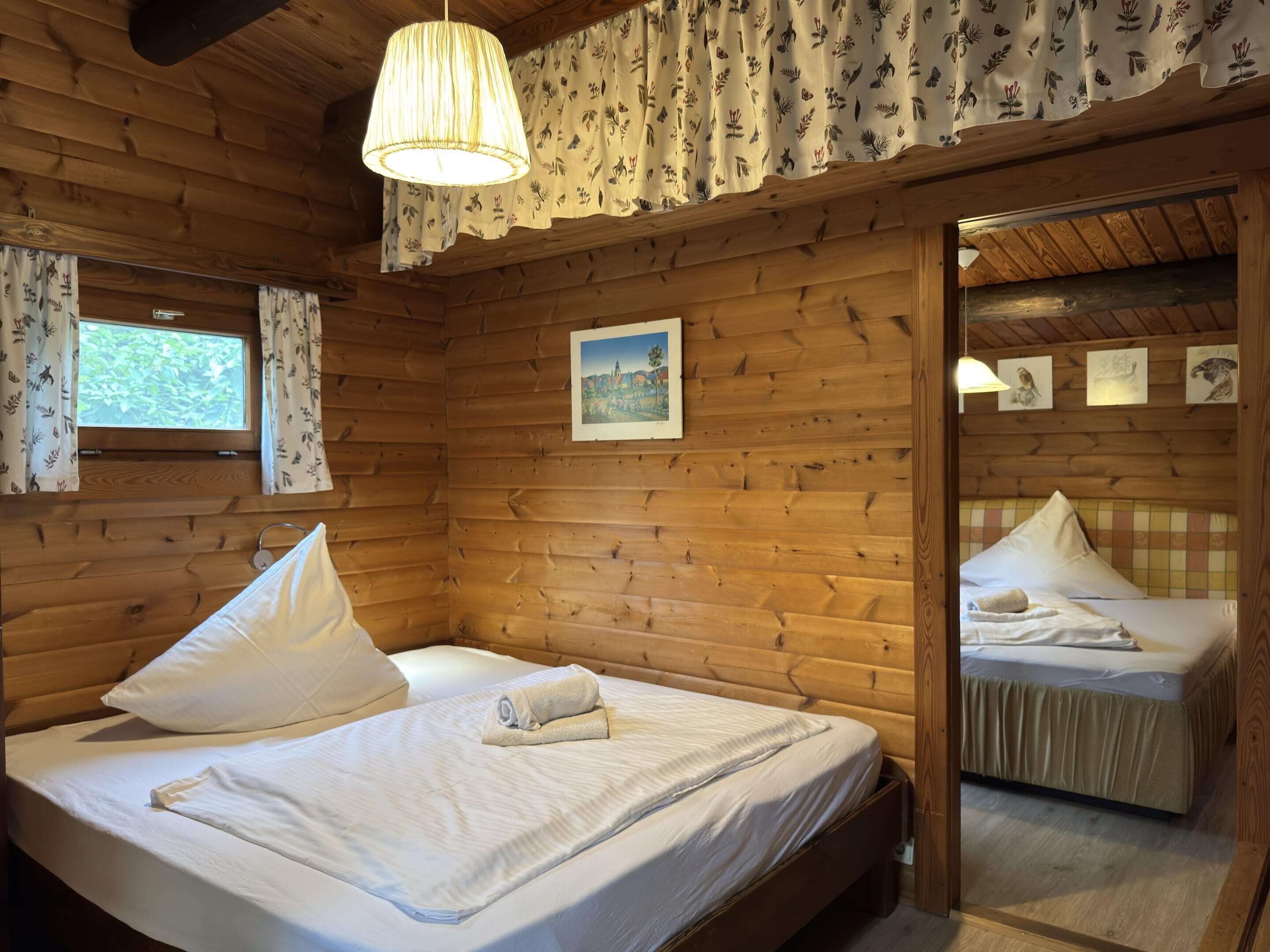 Quarto de cabana de madeira com duas camas, uma janela com cortinas florais e um candeeiro pendente.