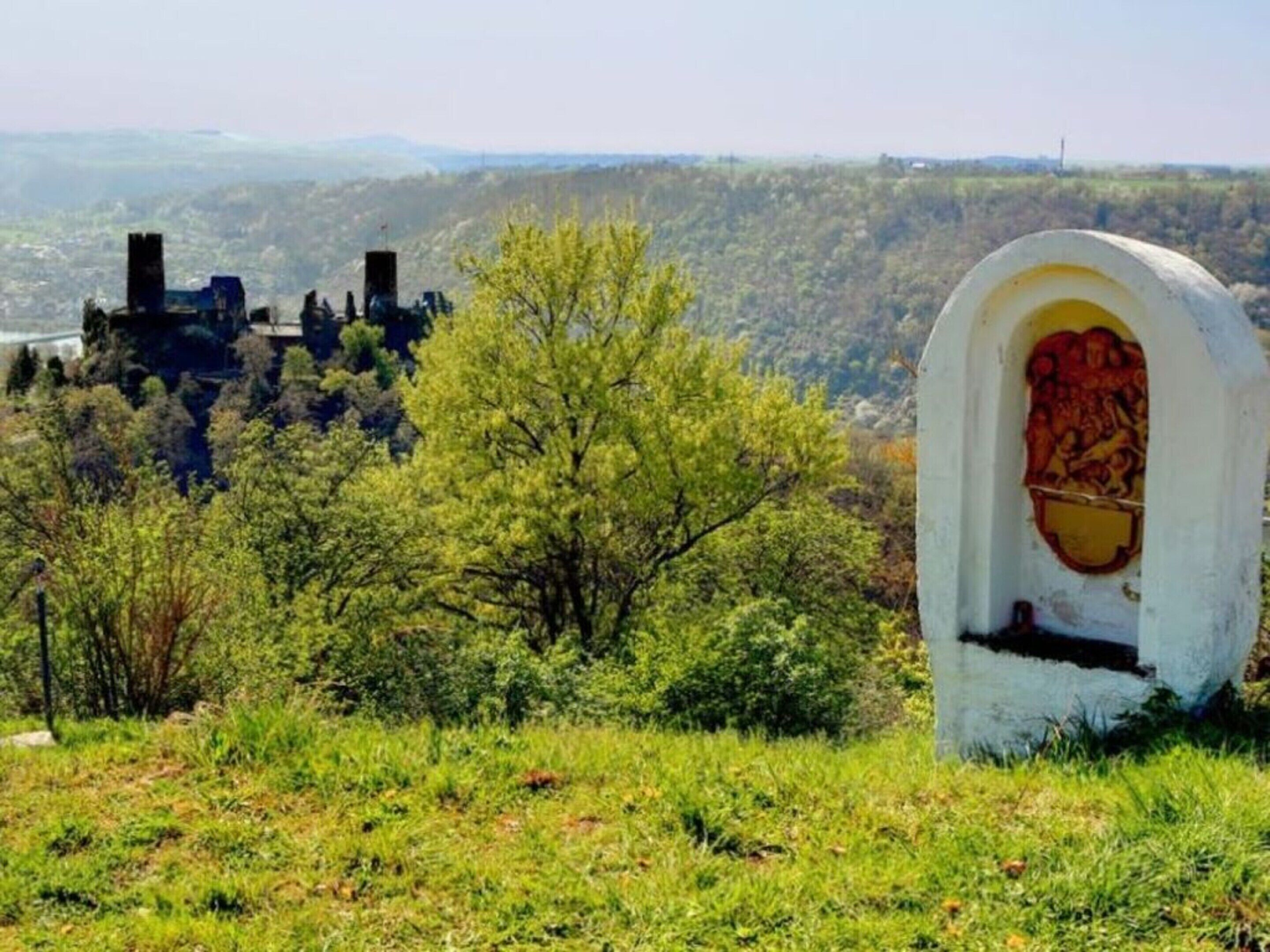 A cemetery with a prominent tombstone featuring a detailed carving, surrounded by lush greenery and a distant castle-like structure.