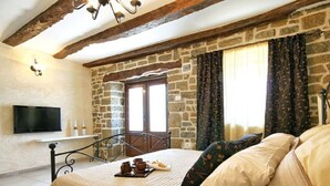 A bedroom with a stone wall, a bed with a beige bedspread, a wooden headboard, a flat-screen TV mounted on the wall, and a window with curtains. - Aurora Comfortable Holiday Residence (Oprtalj)