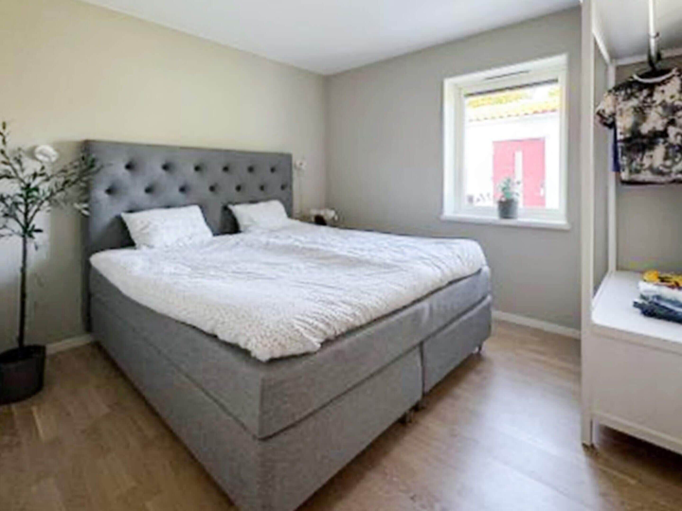 A bedroom with a grey upholstered bed, a grey headboard, and a window with a view of a red building.