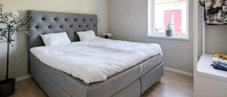 A bedroom with a grey upholstered bed, a grey headboard, and a window with a view of a red building.
