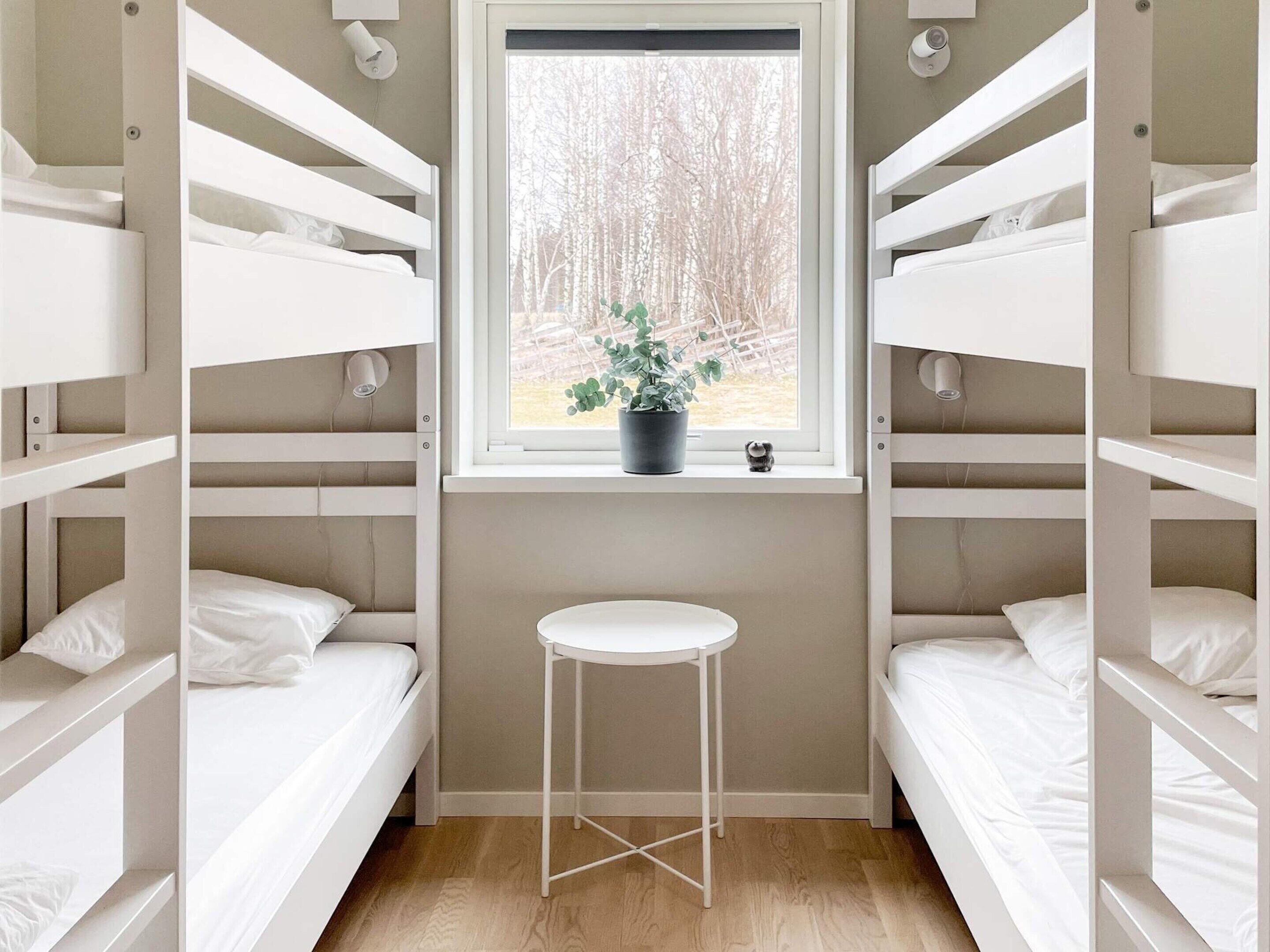 A bunk room with white bunk beds, a small white stool, a window with a view of trees, and a potted plant on the windowsill.