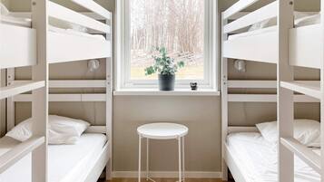 A bunk room with white bunk beds, a small white stool, a window with a view of trees, and a potted plant on the windowsill.