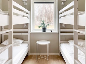 A bunk room with white bunk beds, a small white stool, a window with a view of trees, and a potted plant on the windowsill. - 5 Star Holiday Home in Gotlands Tofta (Gotlands Tofta)