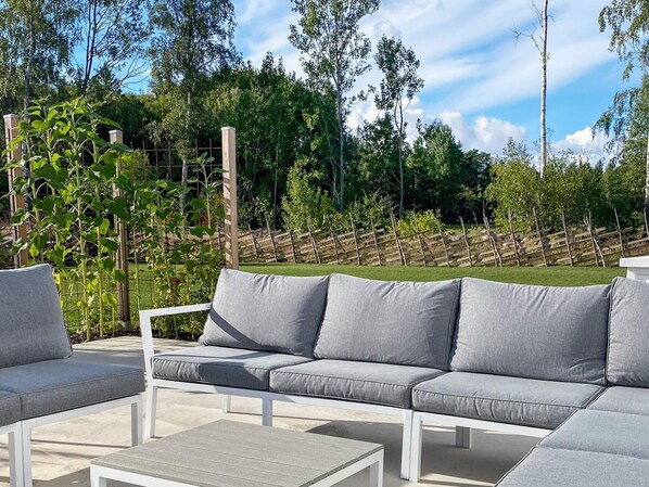 Un área de estar al aire libre con un sofá gris y sillones blancos, rodeada por una valla de madera y follaje.