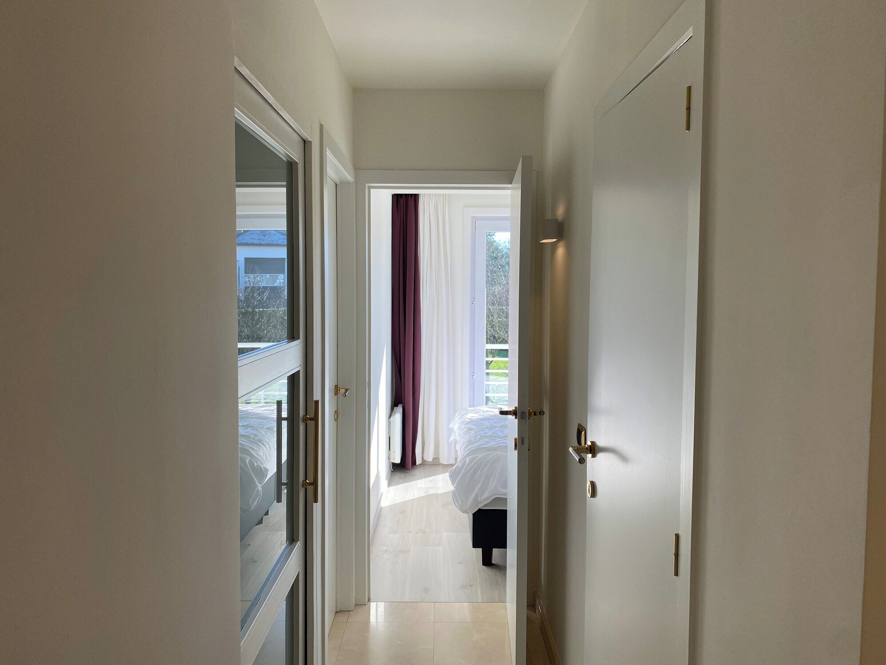 A hallway leading to a bedroom with a bed, a window with curtains, and a view of the outdoors.