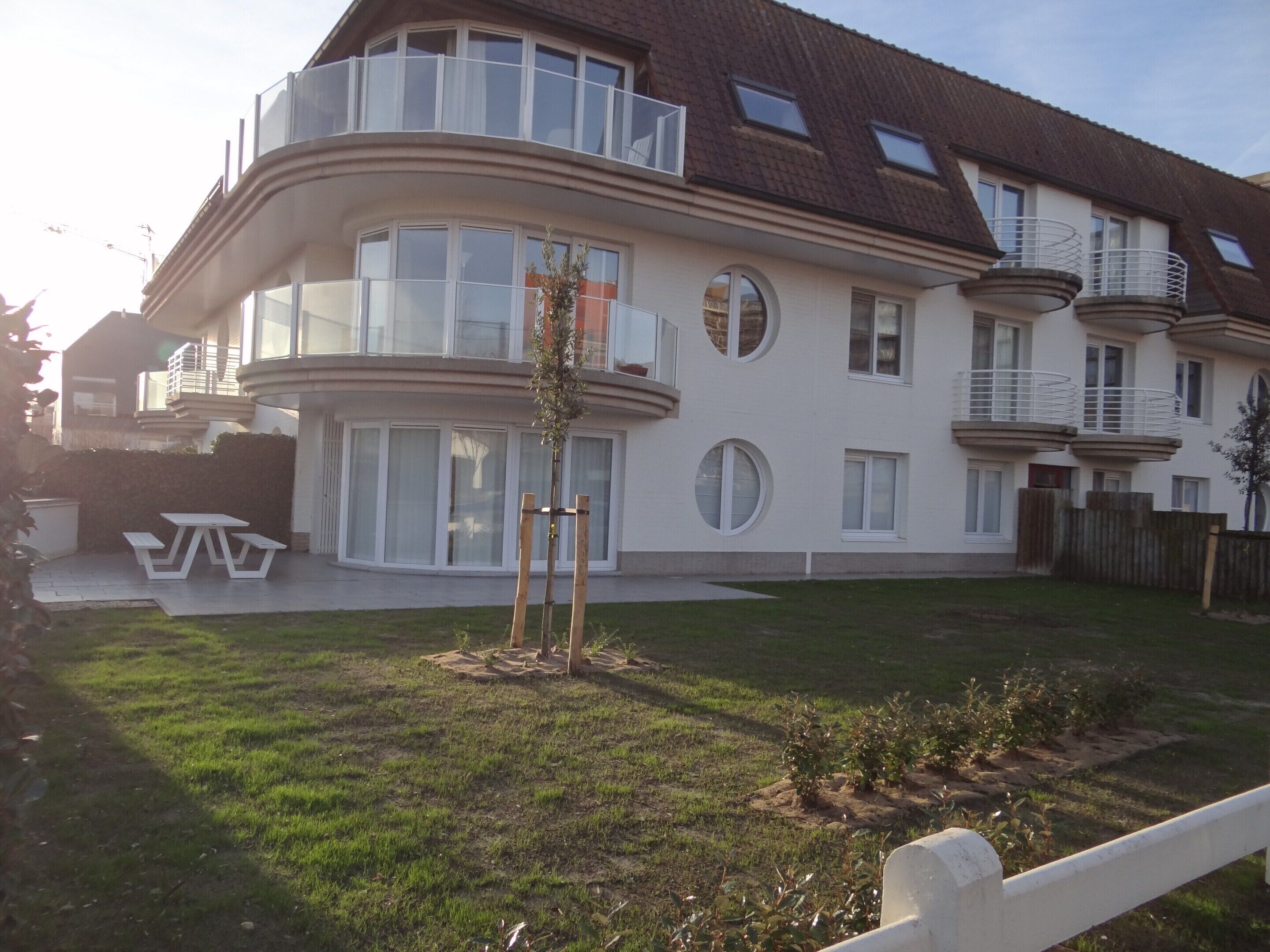 A modern two-story house with a curved balcony, large windows, and a small garden area with a picnic table.