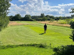 Un campo de golf con una persona de pie en el green, rodeado de árboles y zonas de césped abierto.