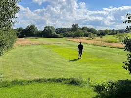 Un campo de golf con una persona de pie en el green, rodeado de árboles y zonas de césped abierto.