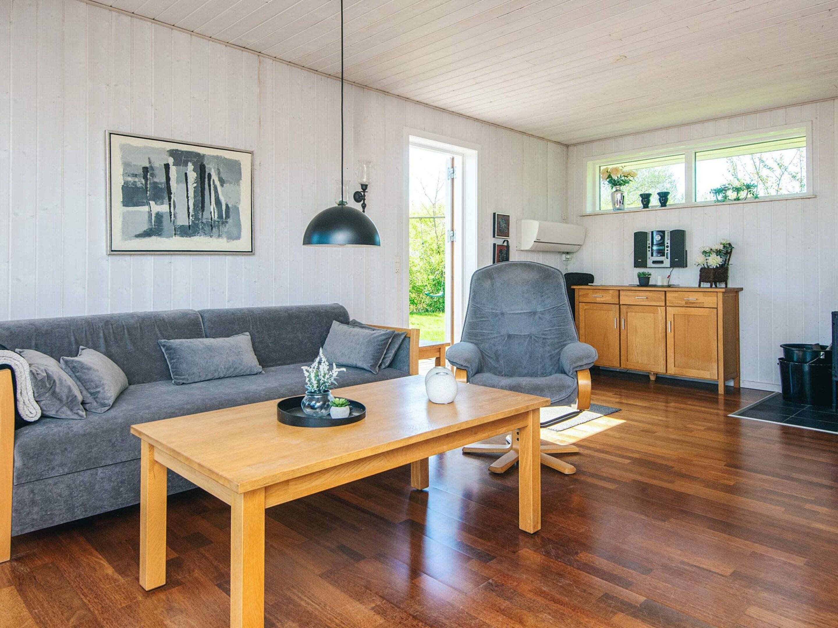 Un salon moderne avec un canapé gris, une table basse en bois et un fauteuil gris. On y trouve également un meuble en bois sur lequel est posée une télévision, ainsi qu’une fenêtre agrémentée de plantes.