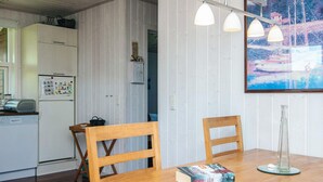 Une table de salle à manger en bois, sur laquelle est posée une pile de livres, deux chaises, un coin cuisine avec un réfrigérateur et un tableau accroché au mur.
