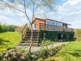 A wooden cabin with a staircase leading to a deck, surrounded by greenery and a clear sky.