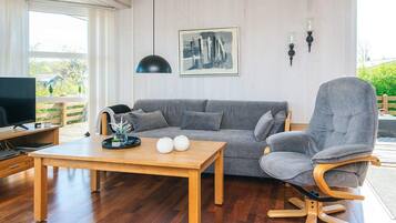A modern living room with a grey sofa, a wooden coffee table, a TV on a wooden stand, and a large window with a view of greenery.
