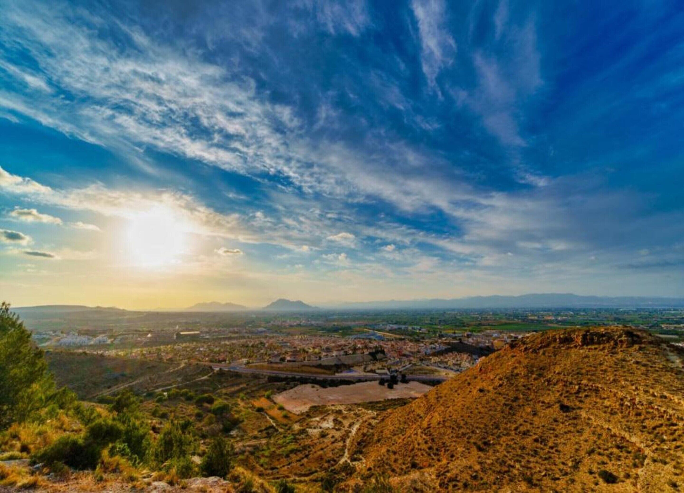A panoramic view of a landscape with a clear sky, distant mountains, and a valley with buildings and greenery.