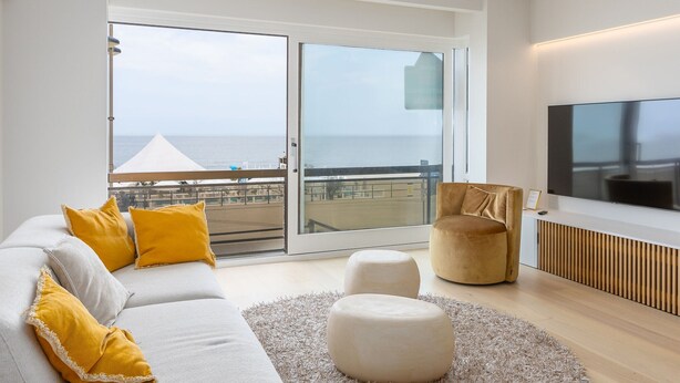 A modern living room with a sofa, ottoman, and armchair, a flat-screen TV, and a balcony with a view of the sea. - Beautiful Apartment With Direct sea View (Middelkerke)