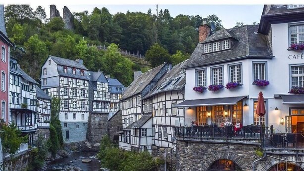 Blick auf ein Flussufer mit traditionellen Fachwerkhäusern, einer Steinbrücke und einem Café mit Außensitzplätzen.