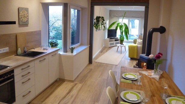 A modern kitchen with a dining area, wooden flooring, and a large window providing natural light.