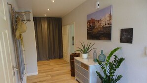 A hallway with wooden flooring, a white dresser with drawers, a potted plant, and a wall-mounted painting of a town.
