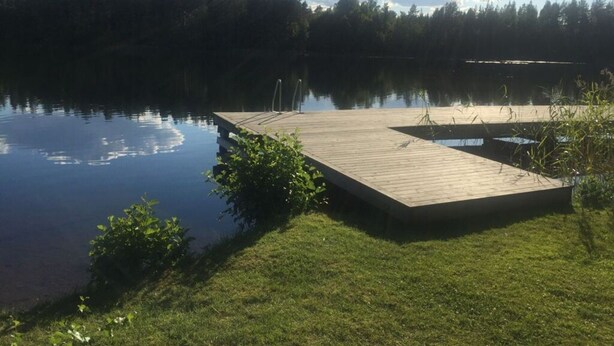 Un molo di legno si protende in un lago calmo, circondato da una vegetazione lussureggiante e una zona boschiva.