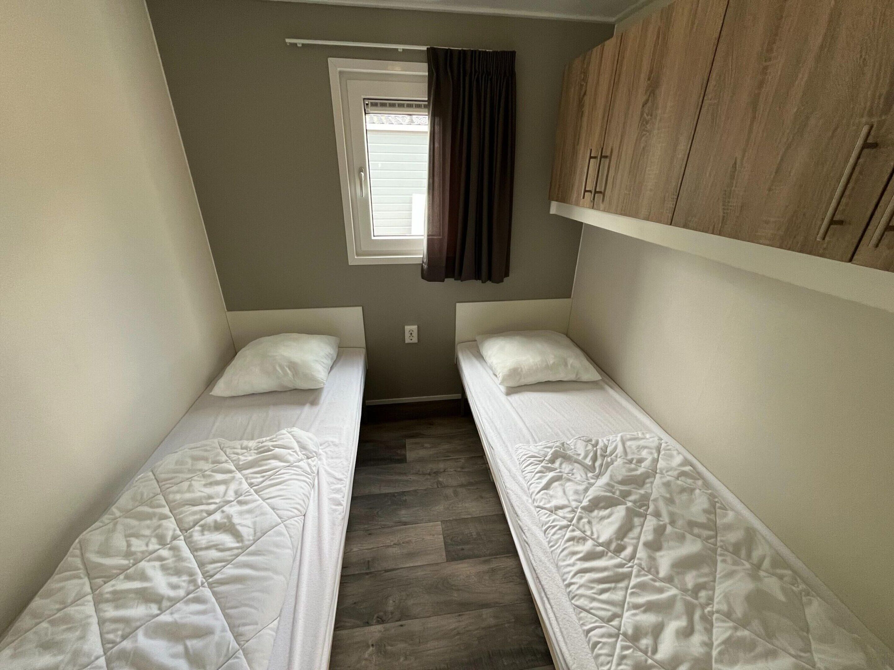 Une petite pièce étroite, avec deux lits simples, une fenêtre avec des rideaux et une armoire en bois.