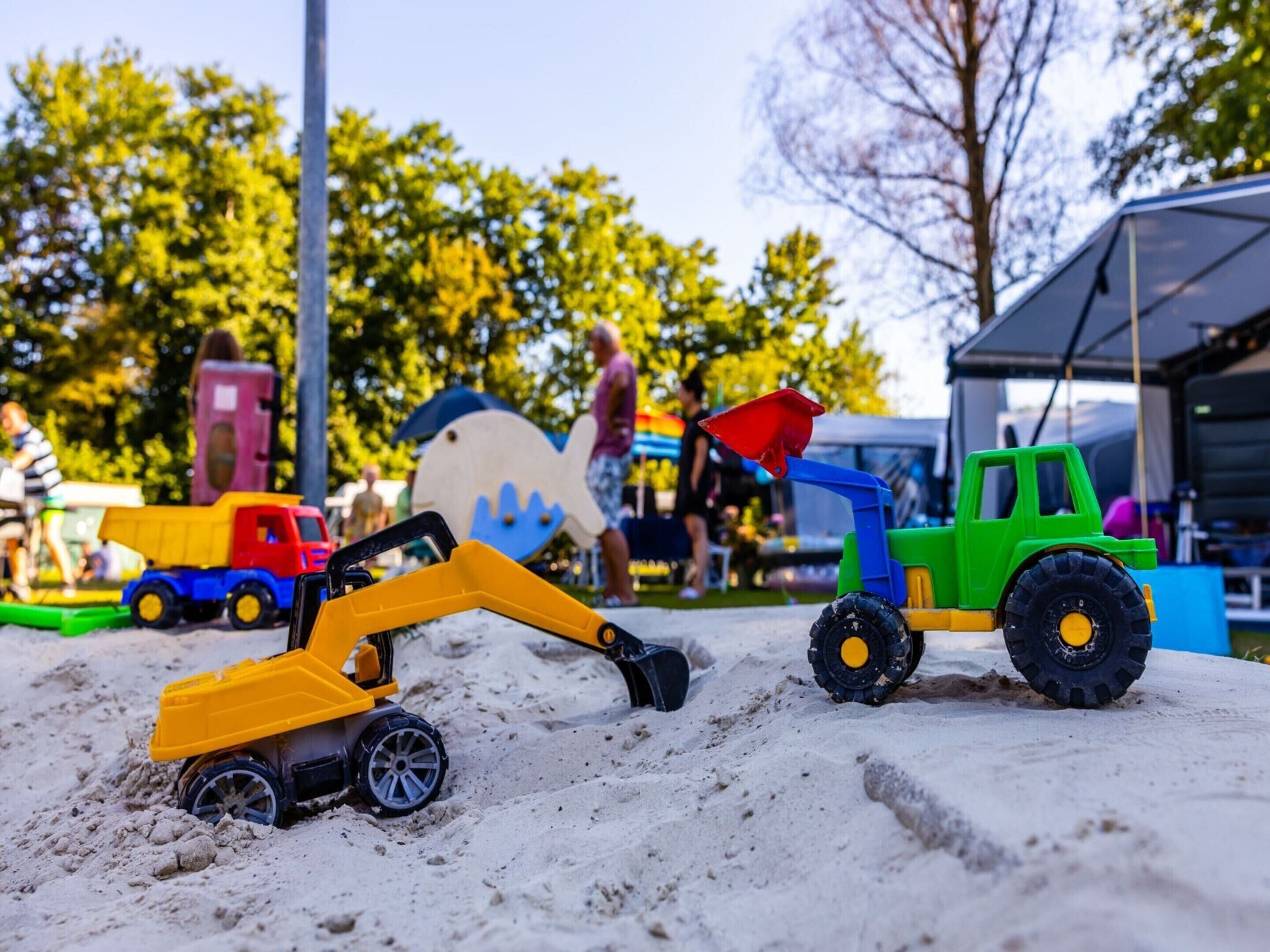 A sandy area with toy vehicles, including a yellow excavator and a green truck.