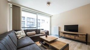 A modern living room with a leather sofa, a wooden coffee table, a TV on a wooden stand, and a large window with a view of buildings. - Moorea City Center Apartment (Middelkerke)